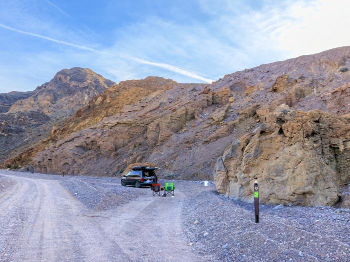 Black camper van with camper chairs in pull-through campsite loop along a gravel dirt road, brown post on ground labelled E-5, canyon walls in the background with blue skies.Site E-5 along Echo Canyon Road.
