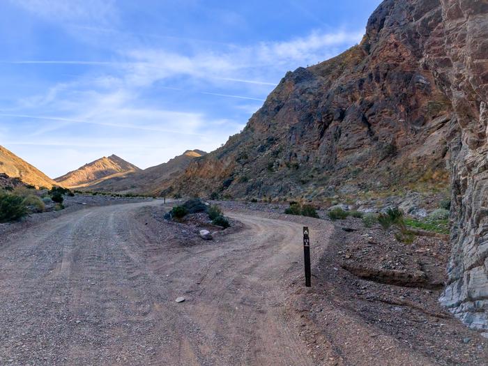 Gravel dirt road with pull-through campsite loop, brown post on ground labelled E-9, mountain backdrop with blue skies.Site E-9 along Echo Canyon Road.
