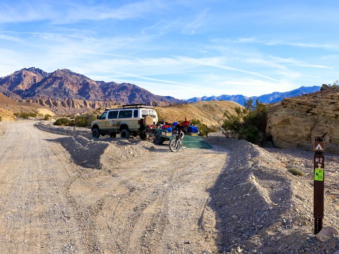 Camper van with trailer in a gravel dirt road with pullout loop and a brown post numbered H1, set against hillsides and mountain backdrop.Site H2 along Hole in the Wall Road