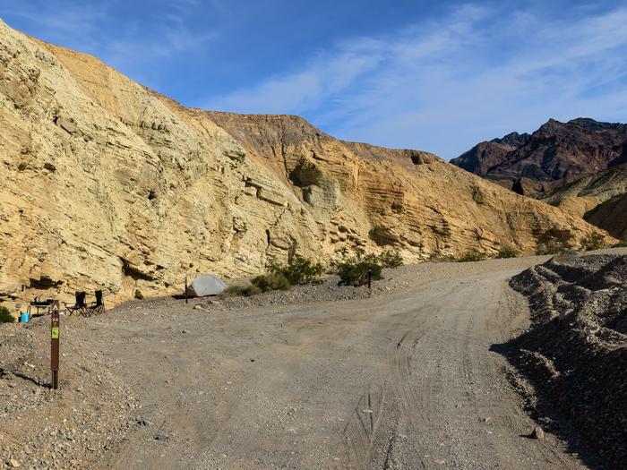 Gravel dirt road with pull-through campsite loop, brown post on ground labelled H4, canyon walls in backdrop with blue skies.Site H4 along Hole in the Wall Road.