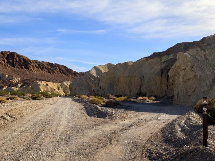 Gravel dirt road with pull-through campsite loop, brown post on ground labelled H5, hillside and mountain backdrop with blue skies.Site H5 along Hole in the Wall Road