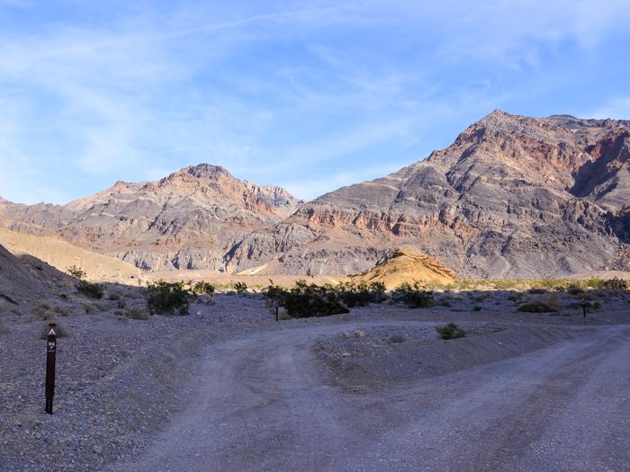 Gravel dirt road with pull-through campsite loop, brown post on ground labelled H6, mountain backdrop with blue skies.Site H6 along Hole in the Wall Road