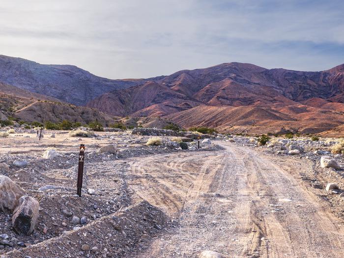 Gravel dirt road with pull-through campsite, brown post on ground labelled M2, hillside backdrop with blue skies.Site M2 along Marble Canyon Road.