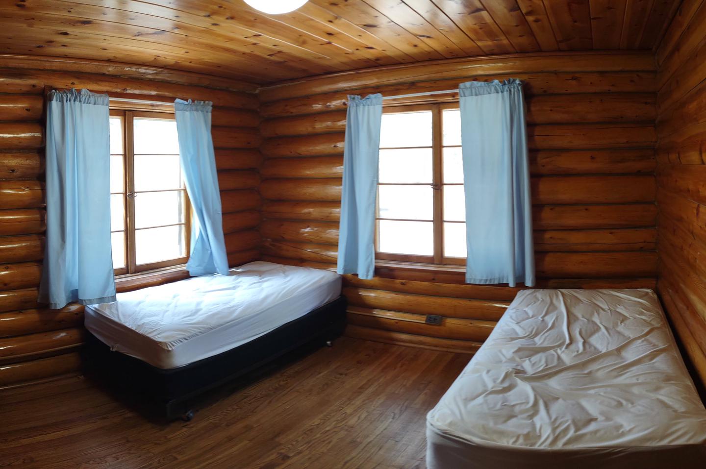 Log cabin bedroom with two twin beds log walls, wood floors and windows with blue drapes. Redfeather Assistant Ranger Cabin Bedroom two. 