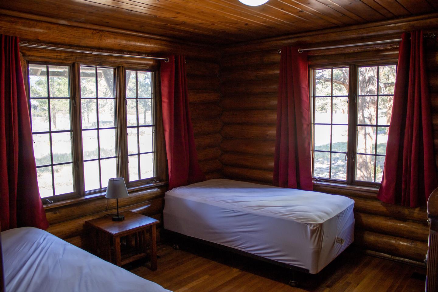 Redfeather Ranger Cabin showing two twin beds, nightstand, lamp, and two large windows with red drapes. Redfeather Ranger Cabin Bedroom one. 