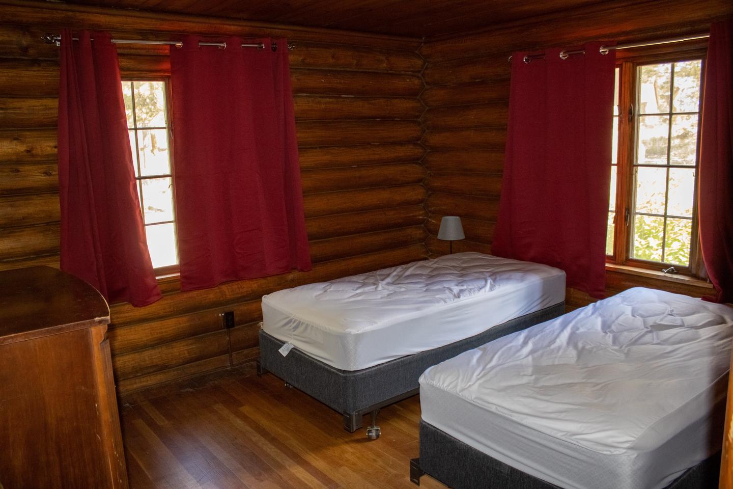 Redfeather Ranger Cabin showing two twin beds, nightstand, lamp, dresser, and two windows with red drapes. Redfeather Ranger Cabin Bedroom two. 