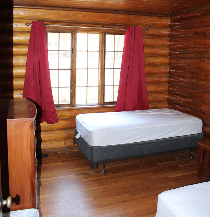 Redfeather Ranger Cabin bedroom showing two twin beds, dresser, nightstand, and window with red drapes. Redfeather Ranger Cabin bedroom three. 