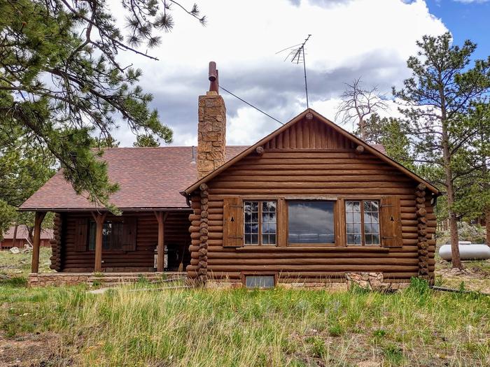 Log cabin with large windows, covered front porch and stone chimney, trees, garage and propane tank in background. Redfeather Ranger Cabin