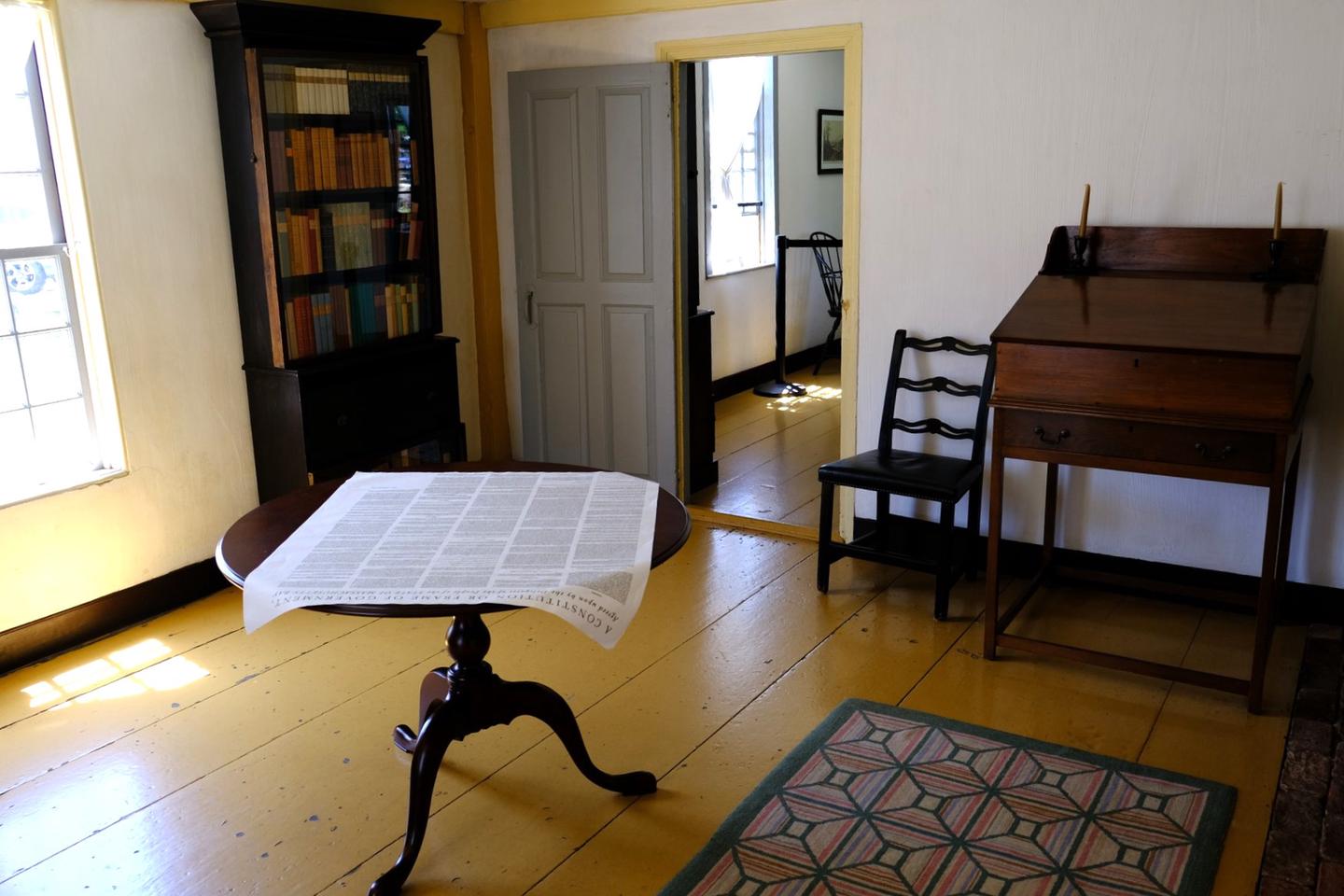 A room with painted yellow floor, circular table in the middle with large broadside on top, standing 18th-century style desk and chair on the right, and bookcase and window on the left. The law office of John Adams inside the John Quincy Adams Birthplace. 