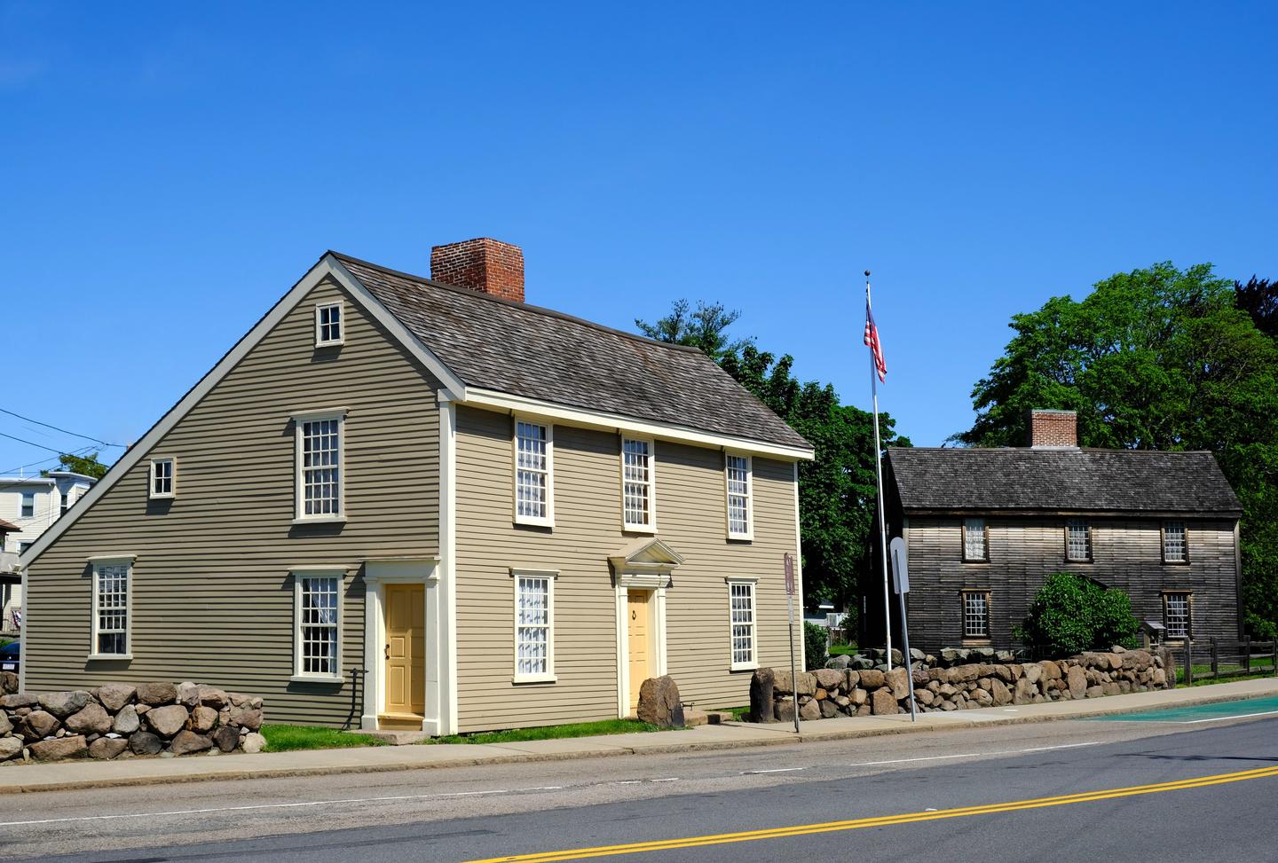 Two New England "saltbox" style homes sit next to each other. Home on left has gray siding, white trim, and beige doors. Home on right has brown wooden siding and is surrounded by trees and shrubs. Between the buildings, a United States flag flies on a flagpole. The Adams Farm at Penn's Hill is home to the John Quincy Adams Birthplace (left) and the John Adams Birthplace (right). 