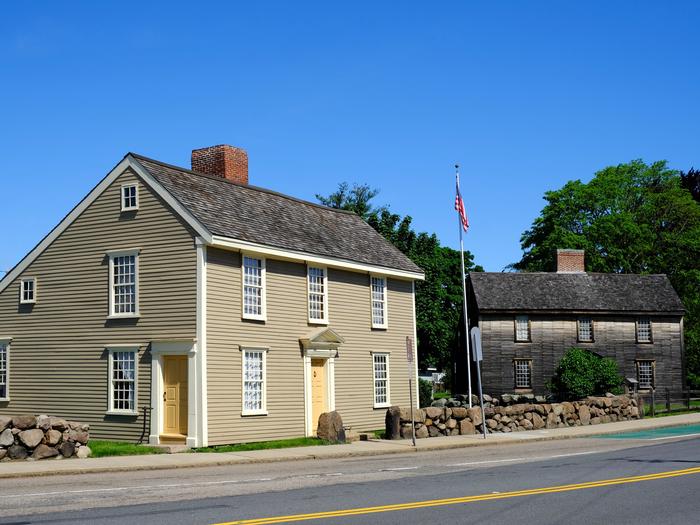 Two New England "saltbox" style homes sit next to each other. Home on left has gray siding, white trim, and beige doors. Home on right has brown wooden siding and is surrounded by trees and shrubs. Between the buildings, a United States flag flies on a flagpole. The Birthplaces of John Adams and John Quincy Adams are part of the Extended Park Tour. 