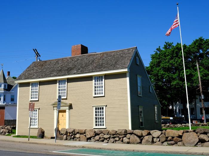 New England "saltbox" style home with light gray siding, windows with white trim, and a yellow door. There is a United States flag flying on a flagpole to the right of the home. The John Quincy Adams Birthplace at the Adams Farm at Penn's Hill is the second stop on the Extended Park Tour. John and Abigail Adams lived in this home with their children in the years before and during the American Revolution. Abigail gave birth to her son and future president, John Quincy Adams, in this home in 1767. 