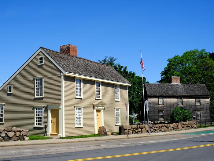 Two New England "saltbox" style homes sit next to each other. Home on left has gray siding, white trim, and beige doors. Home on right has brown wooden siding and is surrounded by trees and shrubs. Between the buildings, a United States flag flies on a flagpole. The John Quincy Adams Birthplace (left) and the John Adams Birthplace (right) at the Adams Farm at Penn's Hill. 