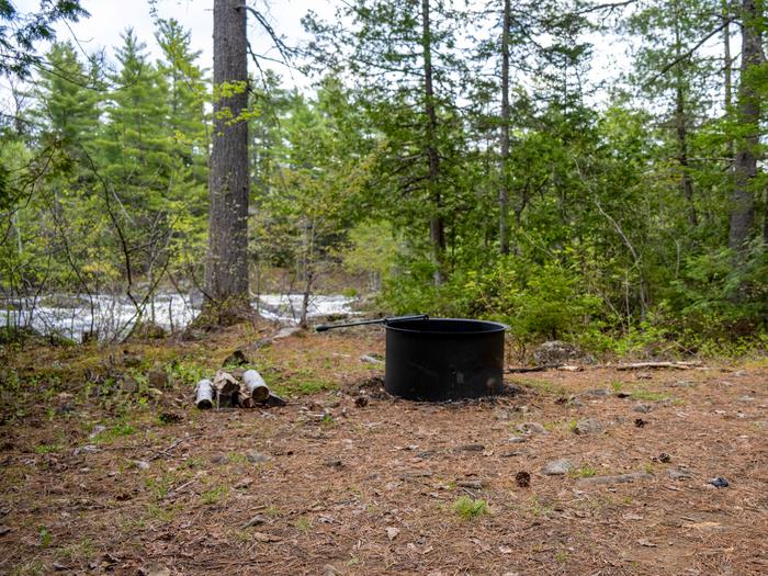 A picture of Pond Pitch Campsite with a black metal firepit and the East Branch of the Penobscot River behind it.Located on the East Branch Penobscot River, Pond Pitch Campsite can be reached by foot or boat.