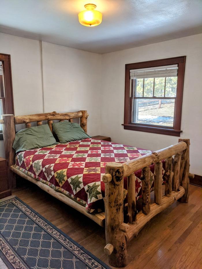 Bedroom with log bed frame, wood floor, white painted walls, four-over-one double hung window and runner rug. Overview of bedroom 1. 