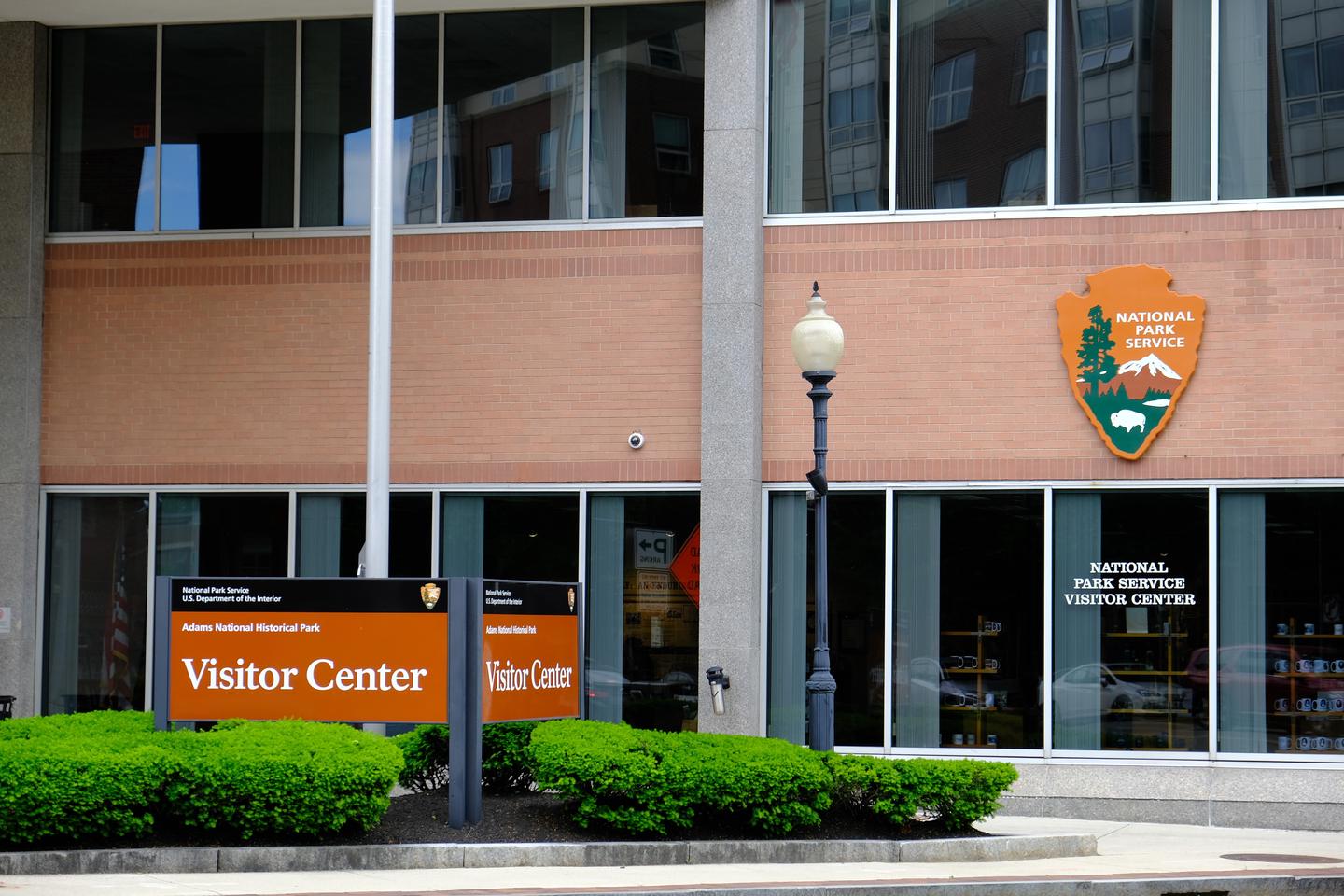 A sign that reads "Visitor Center" stands in front of a brick and glass office building with large windows. A National Park Service arrowhead hangs from the building. The Visitor Center, located at 1250 Hancock St., is a great place to start your day at Adams National Historical Park.