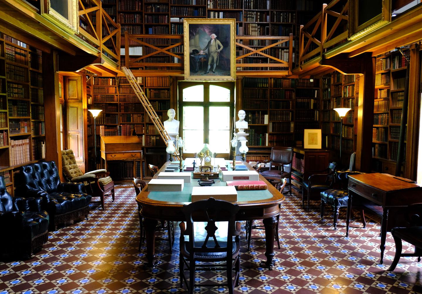 A dark room with tile floors, desks, and chairs. A long table sits at the center. Books on shelves line the walls. The Stone Library at Peace field houses the books of John Quincy Adams and other family mementoes. 