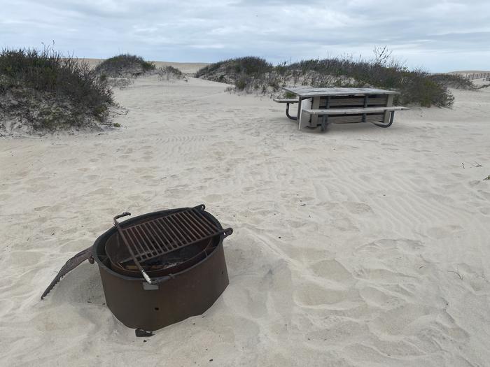 Oceanside site 45 in May.  View is a close-up of black metal fire ring with wooden picnic table nearby on the sand.Oceanside site 45 in May.