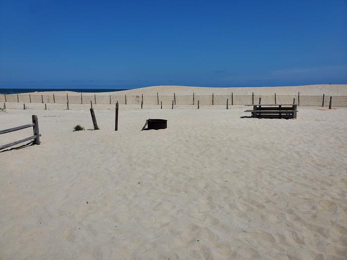 Oceanside site 46 in July.  View of wooden picnic table and black metal fire ring on sand.  Dune line posts are behind the campsite.  Other wooden posts are in the sand nearby including the sign post which has 46 on it.Oceanside site 46 in July.