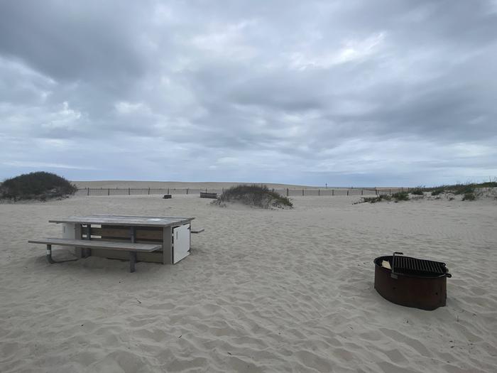 Oceanside site 47 in May 2024.  View of wooden picnic table and black metal fire ring on sand.  There are a couple of dunes behind the campsite that have some brush on it.  Dune line fencing in the background.Oceanside site 47 - May 2024.