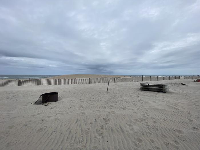 Oceanside site 56 in May.  View of wooden picnic table and black metal fire ring on sand.  Sign post that says 56 on it is in between the picnic table and fire ring.  Dune line posts run along the beach behind the campsite.Oceanside site 56 in May.