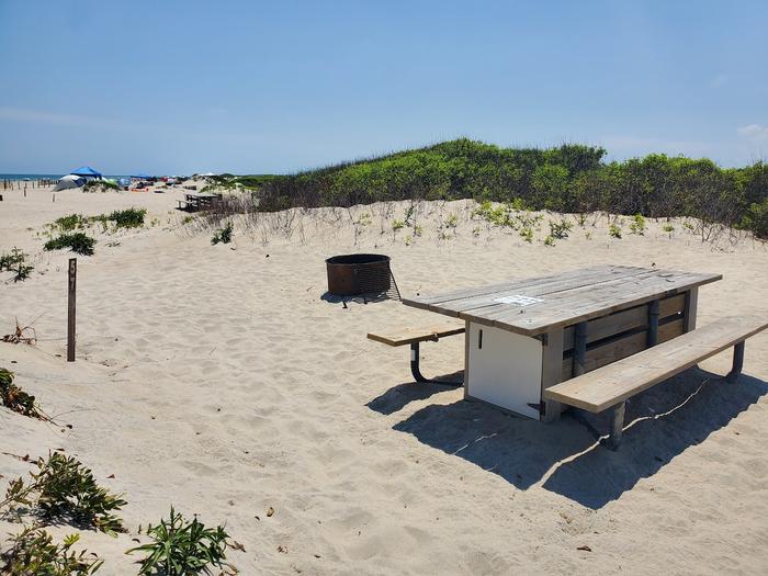 Oceanside site 57 in July.  Close up view of wooden picnic table and black metal fire ring on the sand.  Sign post that says 57 on it is near by.  Other campers are on the horizon.Oceanside site 57 in July.