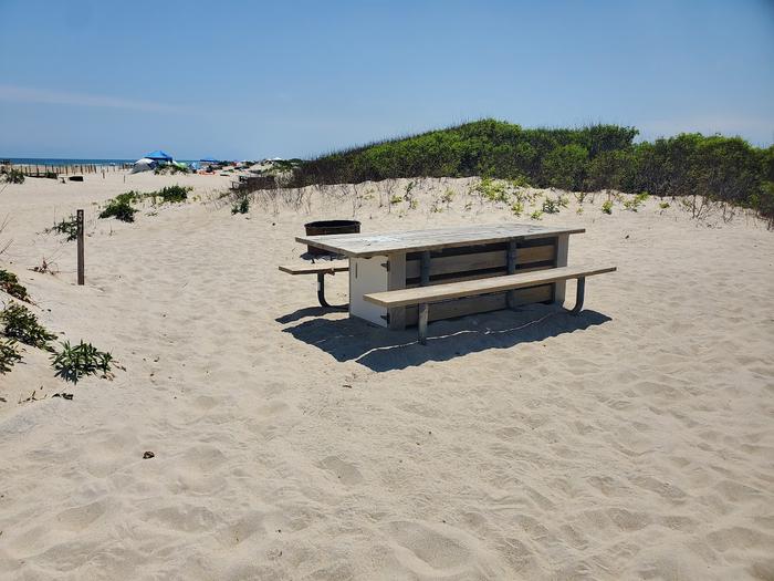 Oceanside site 57 in July.  View of wooden picnic table on the sand with a sign post nearby that says 57 on it.  Black metal fire ring is partially obscured by the picnic table.  Other campers on the horizon.Oceanside site 57 in July.