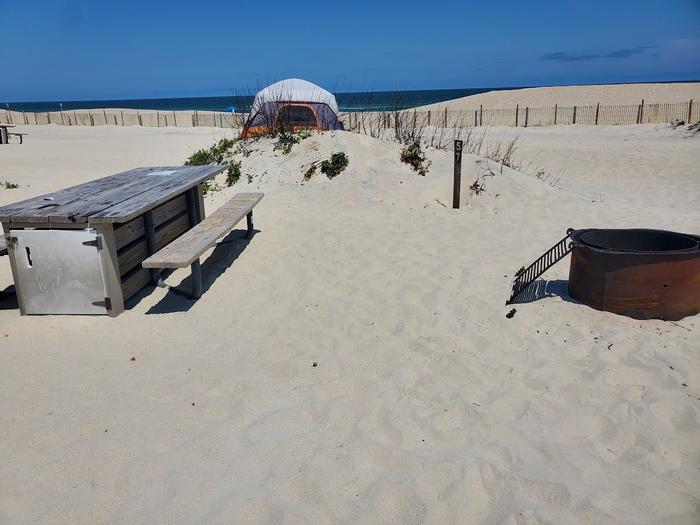 Oceanside site 57 in July.  Close up view of the wooden picnic table and black metal fire ring on the sand.  Sign post nearby that says 57 on it.  Dune line posts run along the beach behind the campsite.  Other campers within view.Oceanside site 57 in July.