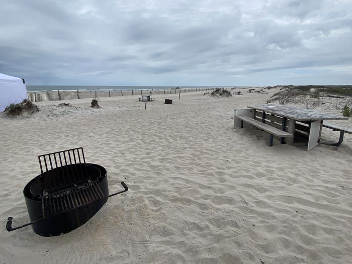 Oceanside site 59 in May.  Close up view of black metal fire ring and wooden picnic table on the sand.  Other campers are within view.  Partial view of the ocean on the horizon.Oceanside site 59 in May.
