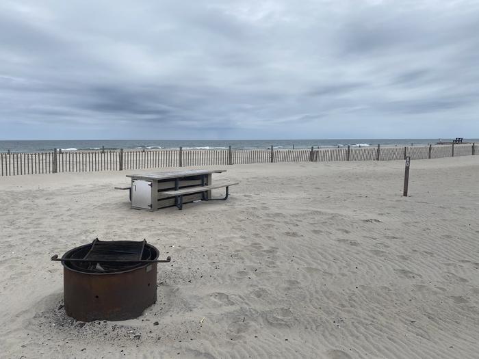 Oceanside site 60 in May.  Close up view of black metal fire ring and wooden picnic table on the sand.  Sign post that says 60 on it is nearby.  Dune line posts run along the beach behind the campsite with a partial view of the ocean on the horizon.Oceanside site 60 in May.