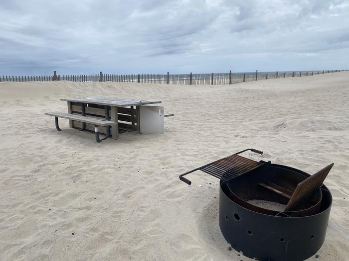 Oceanside site 74 in May.  Close up view of black metal fire ring and wooden picnic table on the sand.  Dune fencing runs along the beach behind the campsite with the ocean front on the horizon.Oceanside site 74 in May.