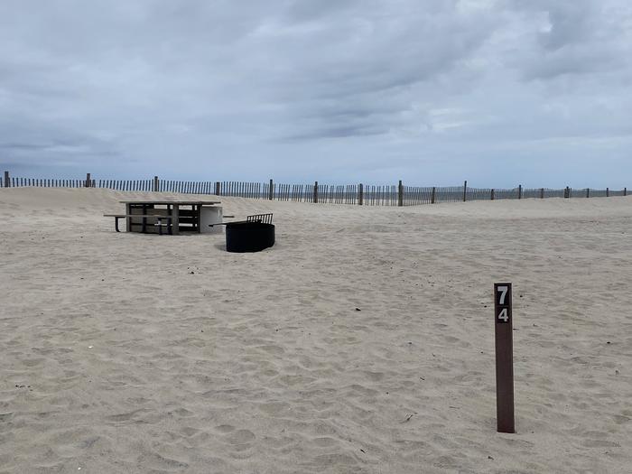Oceanside site 74 in May.  Sign post is closest to the viewer and says 74 on it.  Wooden picnic table and black metal fire ring are behind the campsite.  Dune fencing runs along the beach front behind the table and fire ring. Ocean front on the horizon.Oceanside site 74 in May.