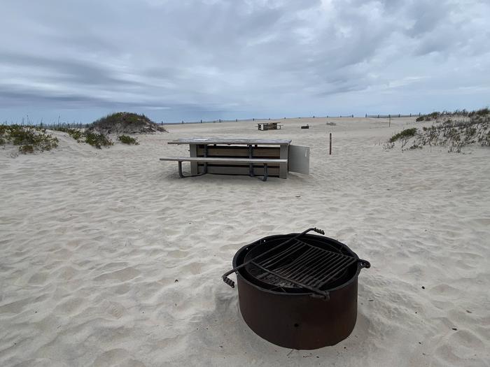 Oceanside site 75 in May.  Close up view of the black metal fire ring with wooden picnic table behind it on the sand.  Sign post nearby says 75 on it.  Other campsites within view.Oceanside site 75 in May.