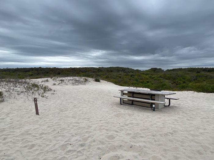 Oceanside site 75 in May.  View of wooden picnic table on the sand.  Sign post nearby says 75 on it.  Black metal fire ring not in the view.Oceanside site 75 in May.