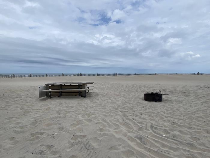 Oceanside site 76 in May.  View of wooden picnic table and black metal fire ring on the sand.  Dune fencing runs along the beach front with the ocean front on the horizon.Oceanside site 76 in May.
