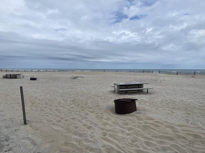 Oceanside site 77 in May.  View of wooden picnic table and black metal fire ring on the sand.  Sign post nearby says 77 on it.  Dune fencing runs along the beach behind the campsite with the ocean on the horizon. Other campsites within view.Oceanside site 77 in May.