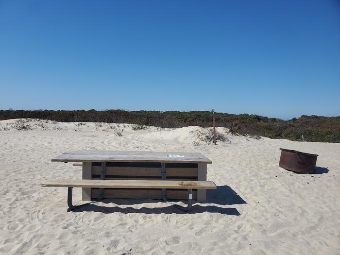 Oceanside site 79 in October.  Close up view of wooden picnic table and black metal fire ring on the sand.  Sign post nearby says 79 on it.  Brush runs along the horizon.Oceanside site 79 in October.