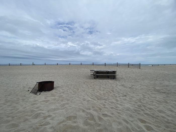 Oceanside site 81 in May.  View of black metal fire ring and wooden picnic table on the sand.  Dune fencing runs along the beach on the horizon.Oceanside site 81 in May.