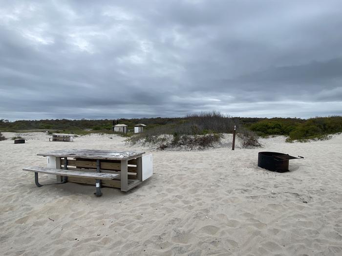Oceanside site 84 in May.  View of the black metal fire ring and wooden picnic table on the sand.  Sign post nearby says 84 on it.  Brush along the horizon.  Other campsites within the view as well as the bathrooms.Oceanside site 84 in May.