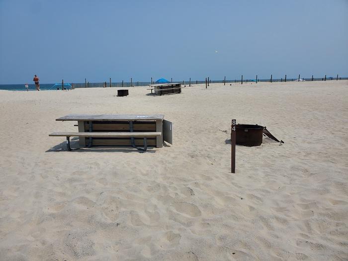 Oceanside site 84 in July.  View of the wooden picnic table and black metal fire ring on the sand.  Sign post in front of the campsite with 84 on it.  Dune fencing runs along the beach with other campsites within the view.Oceanside site 84 in July.