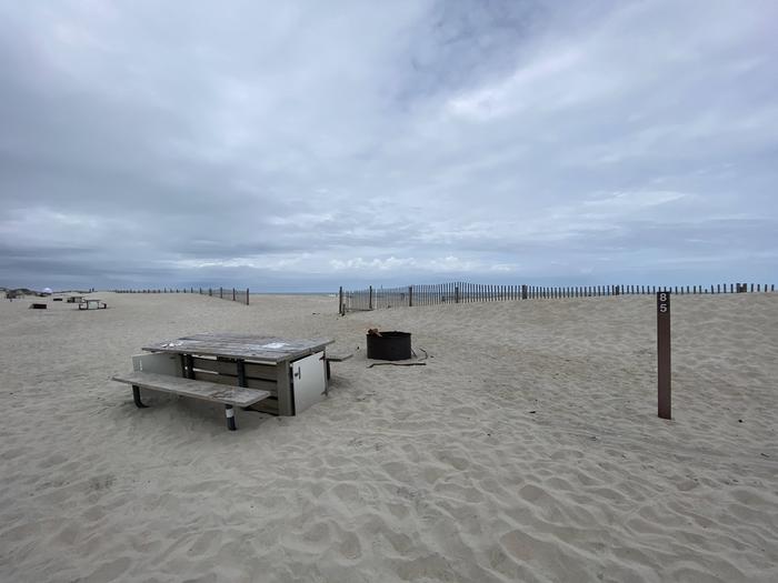 Oceanside site 85 in May.  View of wooden picnic table and black metal fire ring on the sand.  Sign post nearby says 85 on it.  Dune fencing runs along the beach with an entrance leading to the ocean front.Oceanside site 85 in May.