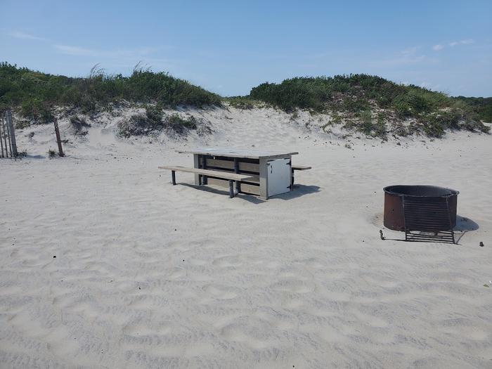 Oceanside site 90 in August.  View of wooden picnic table and black metal fire ring on the sand.  Sign post nearby says 90 on it.  Dune fencing can be seen right at the left corner of the image.  Dune behind the campsite.Oceanside site 90 in August.