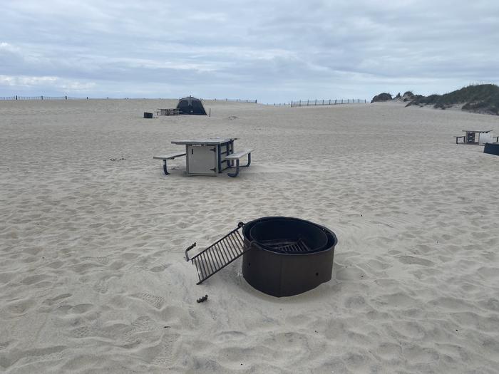 Oceanside site 91 in May.  Close up view of the black metal fire ring on the sand.  Wooden picnic table is behind it.  Other campsites and tents within the view.  Dune fencing runs along the horizon.Oceanside site 91 in May.