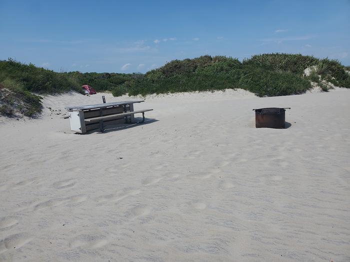 Oceanside site 91 in August.  View of the wooden picnic table and black metal fire ring on the sand.  Dunes run along the horizon with another camper's tent in the view.Oceanside site 91 in August.