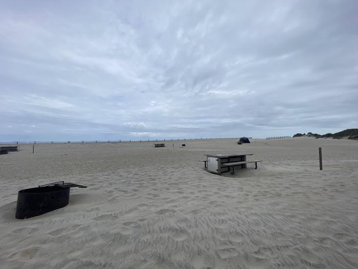 Oceanside site 92 in May.  View of the black metal fire ring and wooden picnic table on the sand.  Sign post nearby says 92 on it.  Dune fencing runs along the beach front on the horizon.Oceanside site 92 in May.