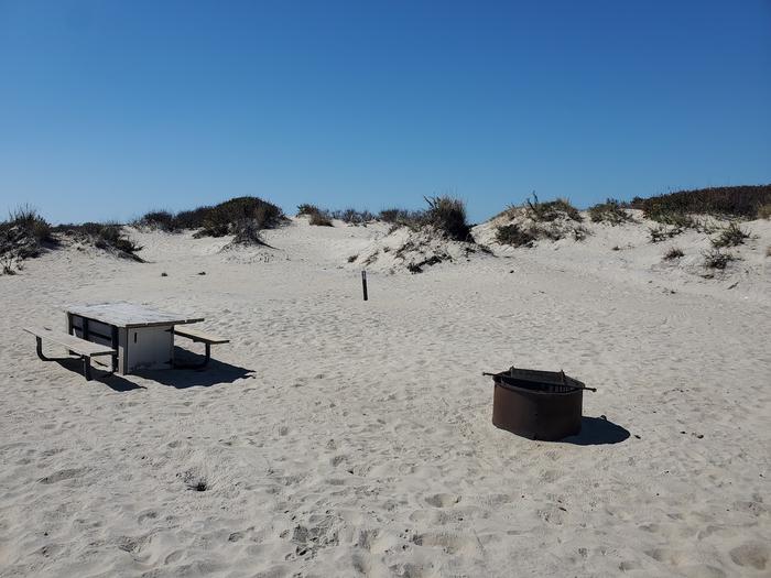 Oceanside site 93 in October 2023.  View of the wooden picnic table and black metal fire ring with a grill on the sand.  Signpost nearby says "93" on it.  Dunes behind the campsite.Oceanside site 93 - October 2023.