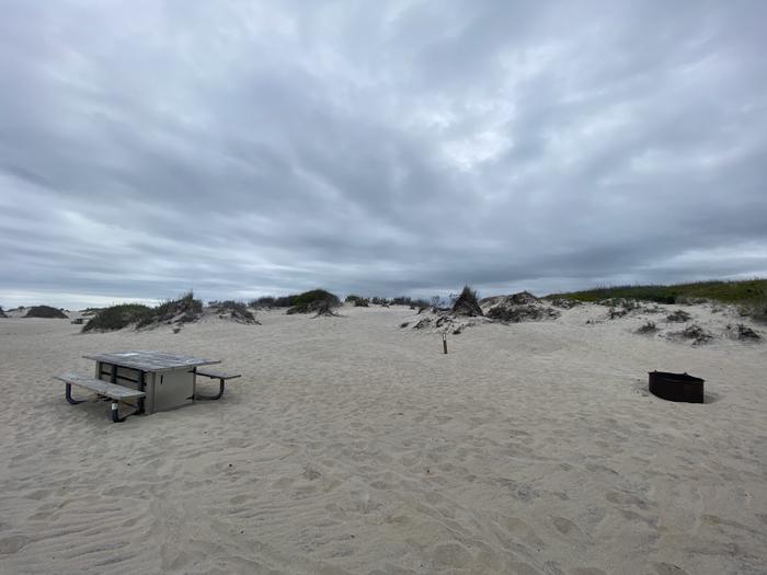 Oceanside site 93 in May 2024.  View of wooden picnic table and black metal fire ring on the sand.  Signpost nearby says "93" on it.  Dunes run along the horizon behind the campsite.Oceanside site 93 - May 2024.