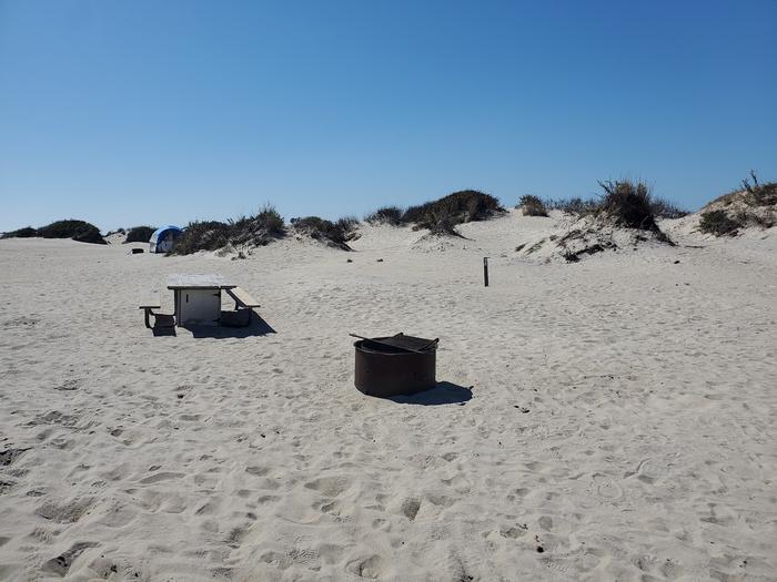 Oceanside site 93 in October 2023.  View of the black metal fire ring and wooden picnic table on the sand.  Signpost nearby that says "93" on it.  Dunes run along the horizon.Oceanside site 93 - October 2023.