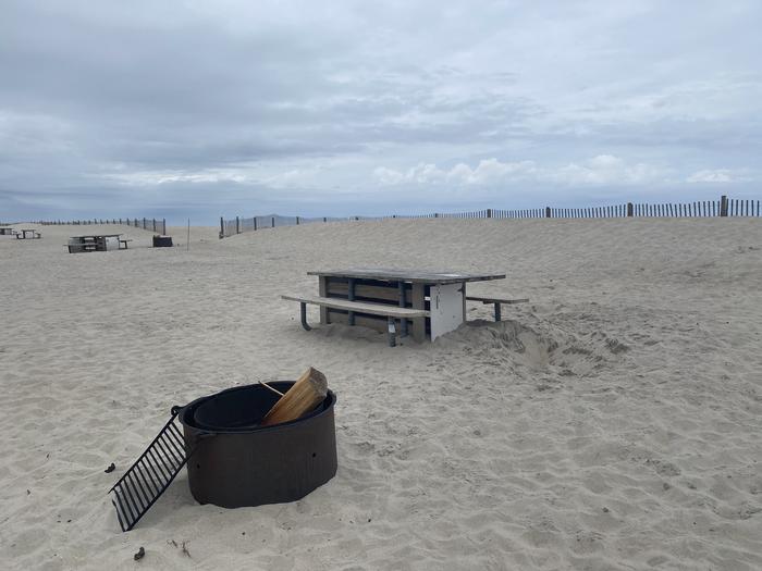 Oceanside site 94 in May.  View of the wooden picnic table and black metal fire ring on the horizon.  There's a piece of partially burned wood sticking out of the fire ring.  Dune fencing runs along the beach front on the horizon.  Other campsites within the view.  Oceanside site 94 in May.
