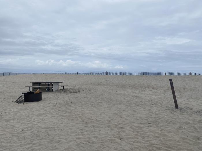 Oceanside site 94 in May.  View of black metal fire ring and wooden picnic table on the sand.  Sign post nearby says 94 on it.  Dune fencing runs along the beach front on the horizon.Oceanside site 94 in May.